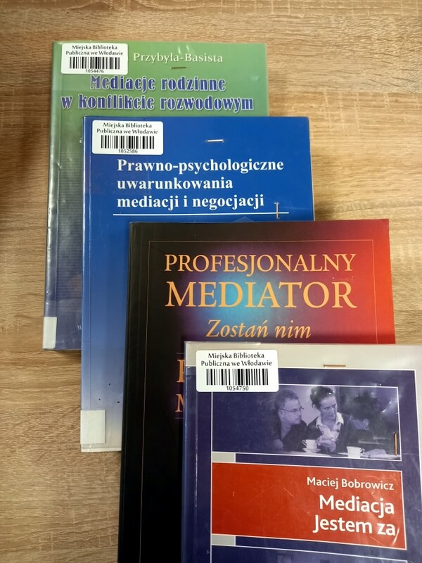Na stole leżą przodem cztery książki dotyczące mediacji, każda kolejna jest przesunięta w ten sposób, że widać tytuł i lewy margines. W lewym górnym rogu okładki trzech książek znajduje się kod kreskowy biblioteki. Tytuły książek od góry: Mediacje rodzinne w konflikcie rozwodowym; Prawno-psychologiczne uwarunkowania mediacji i negocjacji; Profesjonalny mediator. Zostań nim; Mediacja. Jestem za.