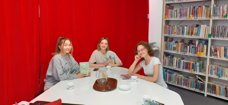 Przy białym, dużym stole siedzą trzy dziewczyny. Każda trzyma w dłoni ulubioną książkę i z uśmiechem spogląda w obiektyw aparatu. W tle z prawej strony stoją wysokie regały biblioteczne z kolorowymi książkami młodzieżowymi. Z lewej strony wisi jaskrawa, czerwona kotara. Na stole stoją filiżanki z herbatą. Brązowa tacka z pustym dzbankiem.