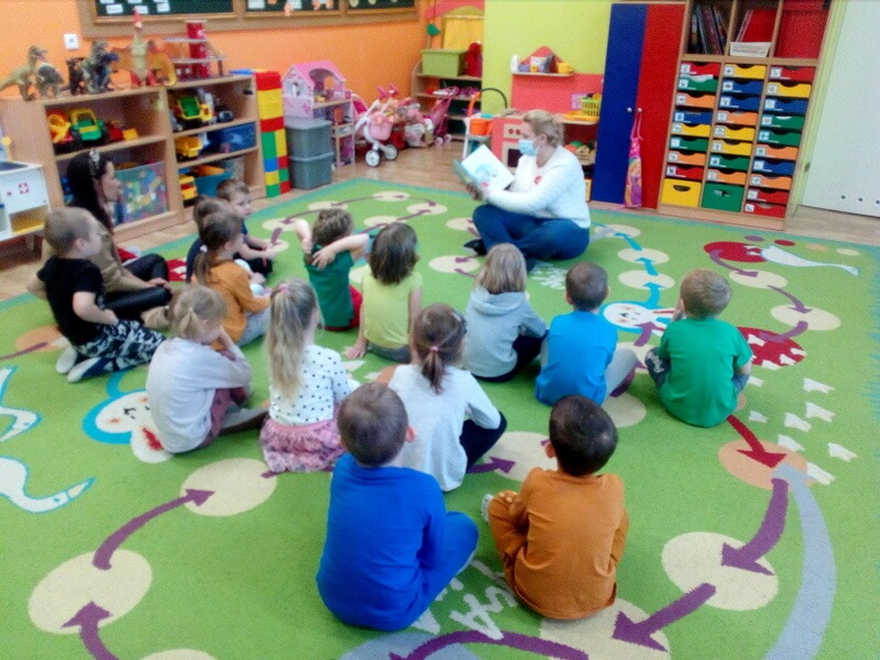 Prowadząca siedzi na kolorowym dywanie i czyta książkę. Tło stanowią mebelki i zabawki dziecięce. Przed nią siedzą dzieci i wychowawczyni, skierowane twarzami do czytającej, a tyłem do osoby, która fotografuje spotkanie.