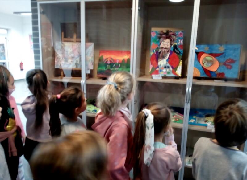 Grupa dzieci skierowana plecami do obiektywu. Dzieci wpatrują się w kolorowe obrazki, które są wyeksponowane w przeszklonej gablocie.