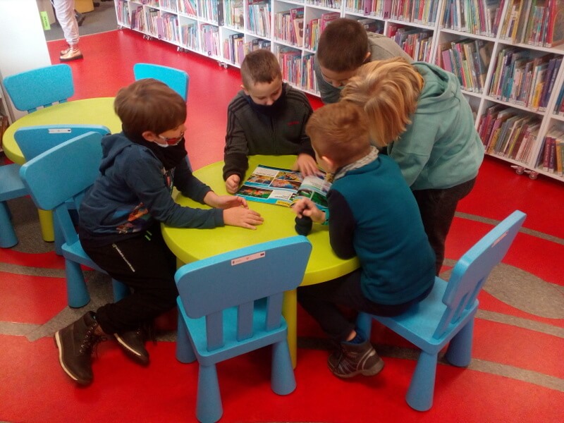 Kadr lekko z góry. Pięciu chłopców siedzi na niebieskich krzesełkach przy zielonym okrągłym stoliku i pochyla się nad czasopismem. Tło zdjęcia stanowi czerwona podłoga Oddziału dla Dzieci oraz widoczny długi, niski regał z książkami dla dzieci.