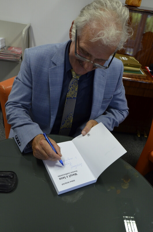 Przy stoliku siedzi pisarz Aldon Dzięcioł i podpisuje swoją książkę.