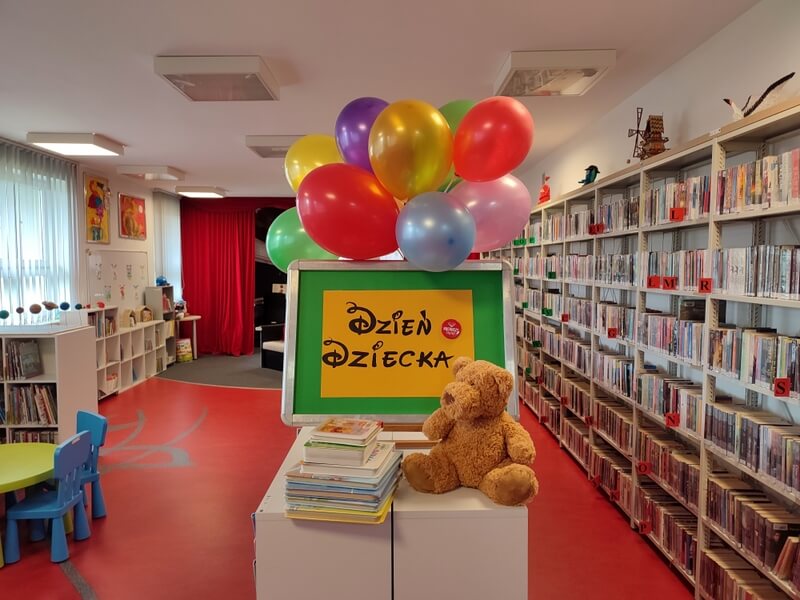 Na niskim regale z książkami jest wyeksponowany napis Dzień Dziecka na żółto-zielonym tle. Wokół niego, są umocowane kolorowe balony. Przed ekspozycją z prawej strony siedzi pluszowy miś, z lewej stos ułożonych książek. Tło stanowi wystrój biblioteki w barwach czerwono – czarnych, mebelki dla dzieci i regały z książkami.