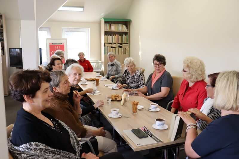 Grupa kobiet siedzi przy stole. Piją kawę i jedzą ciastka. Jedna z Pań pokazuje książki. Wszystkie są pogodne, radosne i uśmiechnięte. W tle duży kolorowy plakat z logo DKK.