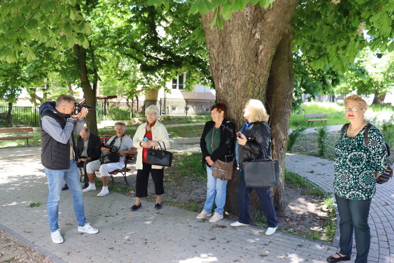 Park. Prowadzący robi zdjęcie dwóm uczestniczkom stoącym pod drzewem. Kilku uczestników przygląda się robieniu zdjęcia. 