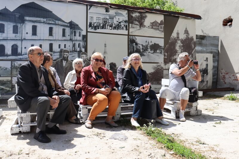 Grupa uczestników siedzi na dworze na drewnianych paletach. W tle, na ścianie, wielkie zdjęcia starej Włodawy. 