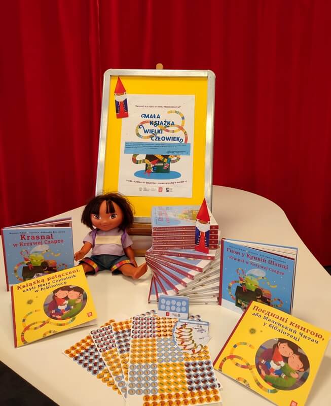 Na pierwszym planie są wyeksponowane książki dla dzieci wraz z naklejkami i kartami Młodego Czytelnika. Między książkami siedzi lalka. Na czerwonym tle znajduje się plakat promujący akcję Mała Książka – Wielki Człowiek.