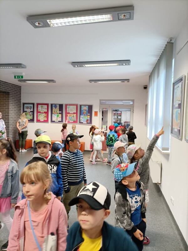 Galeria na Piętrze, grupa dzieci w wieku 6 lat ogląda wystawę eksponowaną w antyramach zawieszonych na ścianach. Pokazują sobie paluszkami ulubione obrazki.