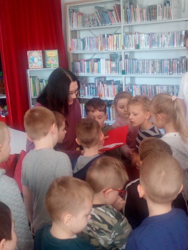 Oddział dla Dzieci, na pierwszym planie grupa przedszkolaków skupiona wokół prowadzącej zajęcia (J. Orzeszko). Uśmiechnięte dzieci i prowadząca oglądają wyszywaną książeczkę. W tle wysokie regały z kolorowymi książkami, fragment czerwonej kotary i szary regał z książkami w języku ukraińskim.