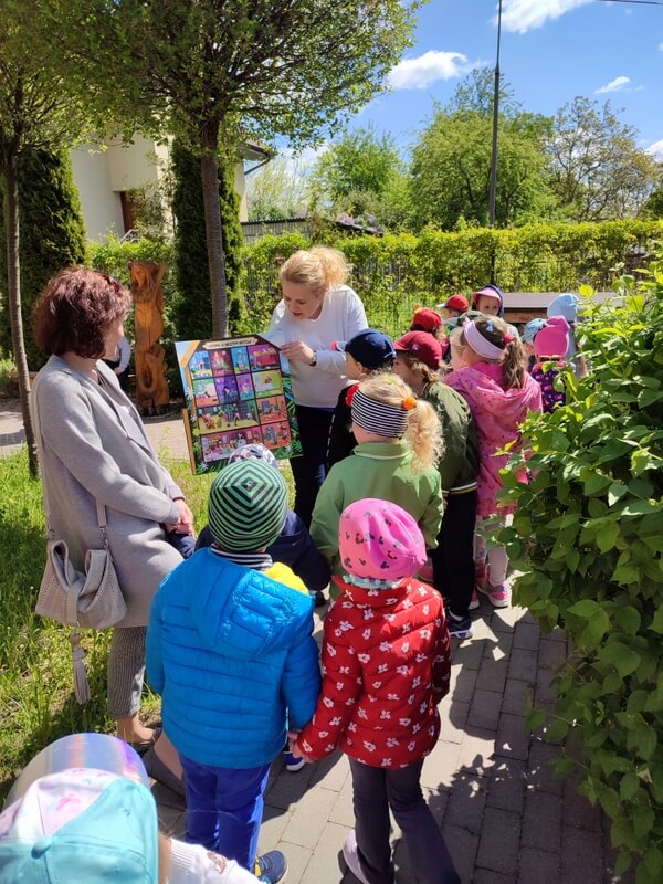 Wokół prowadzącej, która prezentuje kolorową ilustrację stoi grupa dzieci wraz z wychowawczynią. Tło stanowi roślinność w ogrodzie.