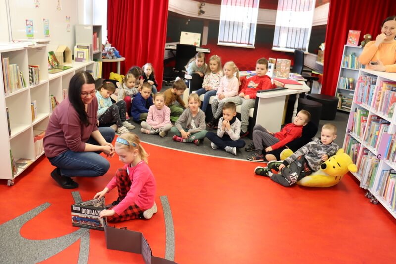 Oddział dla Dzieci. Pierwszy plan czerwona podłoga pomieszczenia, na której stoi rozłożona książka. Przy książce przykuca dziewczynka i prowadząca spotkanie (J. Orzeszko). W tle grupa dzieci siedzi przy białym stole, na szarej wykładzinie oraz na pluszakowych siedziskach dziecięcych. Przyglądają się z zainteresowaniem rozkładanej książce. Po bokach stoją niskie regały z kolorowymi książkami.