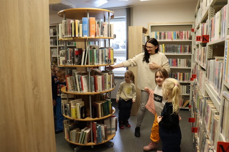 Grupa dzieci zwiedza Wypożyczalnię dla Dorosłych. Dookoła dzieci są wysokie regały w kolorze jasnego drewna, pełne książek. W centrum prowadząca pokazuje obrotowy regał z książkami.
