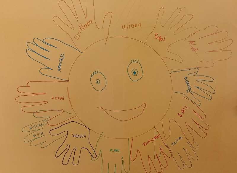 Żółty karton ze wspólną pracą uczestników: uśmiechnięte słońce, którego promienie tworzą odrysowane dłonie dzieci i młodzieży, każda dłoń podpisana imieniem właściciela