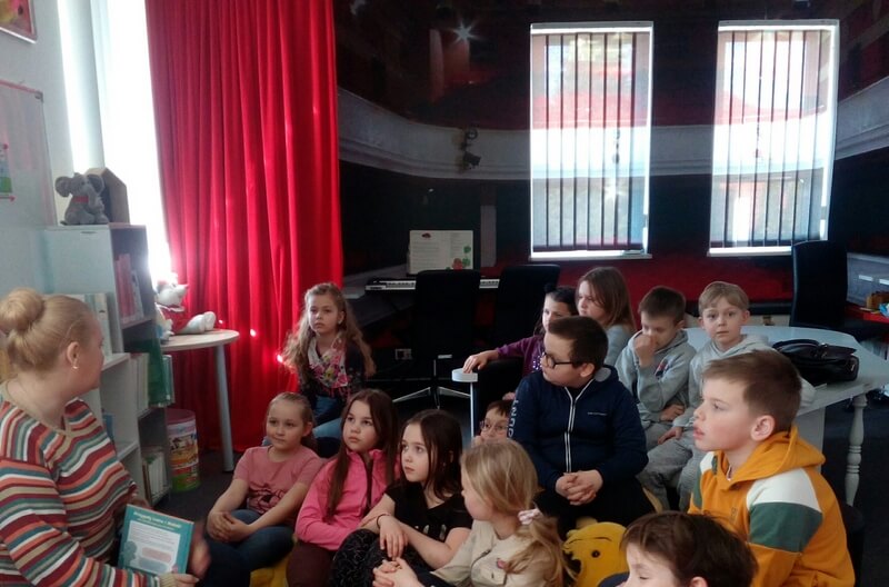 Po lewej stronie siedzi prowadząca zajęcia z książką w dłoni, skierowana do grupy dzieci. Z prawej strony siedzi grupa dzieci, skierowana twarzami do prowadzącej. Tło stanowi wystrój biblioteki w kolorach czerwono- czarnych.