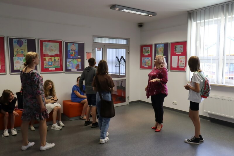 Grupa ludzi (dzieci i dorośli) ogląda wystawę XXI PFTD prac litrackich i plastycznych (prace w antyramach wiszą na ścianach).  