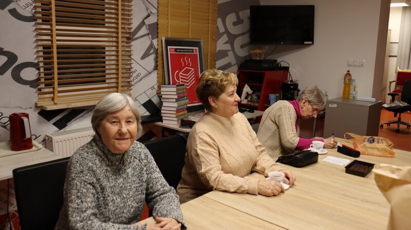 Trzy kobiety siedzące przy stole. Za nimi plakat DKK