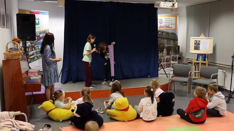 Grupa dzieci siedzi na matach przed sceną. Na scenie dwoje dzieci trzyma karton z rysunkiem. 