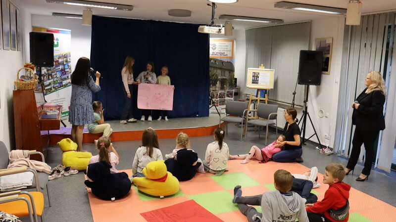Dzieci siedzą na matach przed sceną, na scenie stoją trzy dziewczynki trzymając karton z rysunkiem. 