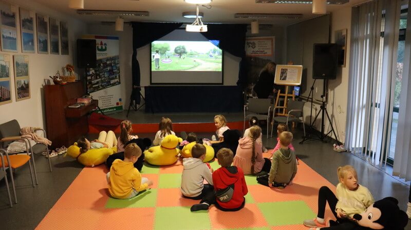 Grupa dzieci siedzi na matach przed sceną. Scena stylizowana na kino, za sceną ekran z kadrem z filmu. 