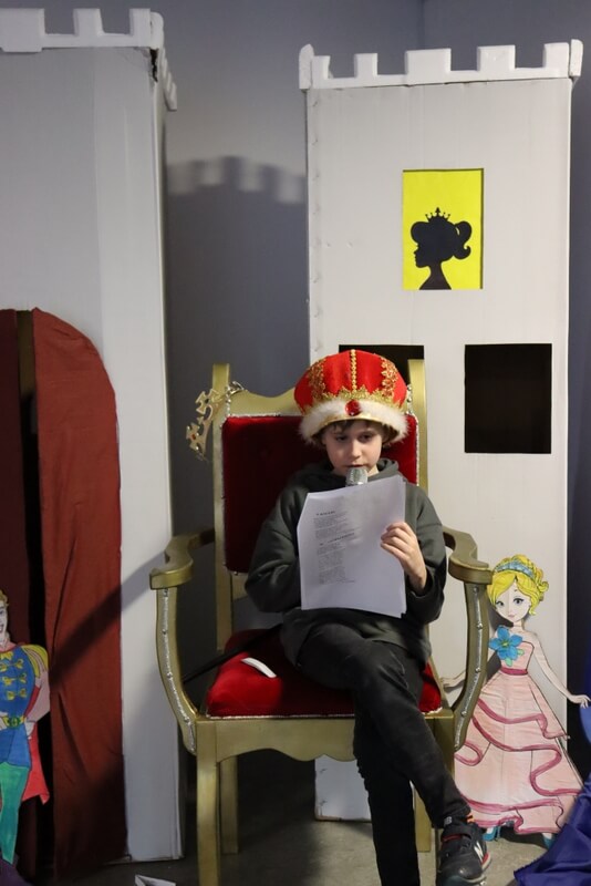 Dziewczynka w koronie siedząca na tronie i czytająca tekst z kartki. Za nią zamkowe wieże wykonane z kartonu. 