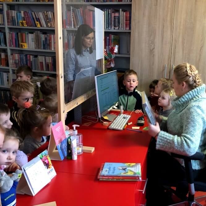 Po prawej stronie siedzi bibliotekarka za biurkiem i wypożycza dzieciom książki, które stoją wokół stołu wraz z wychowawczynią.