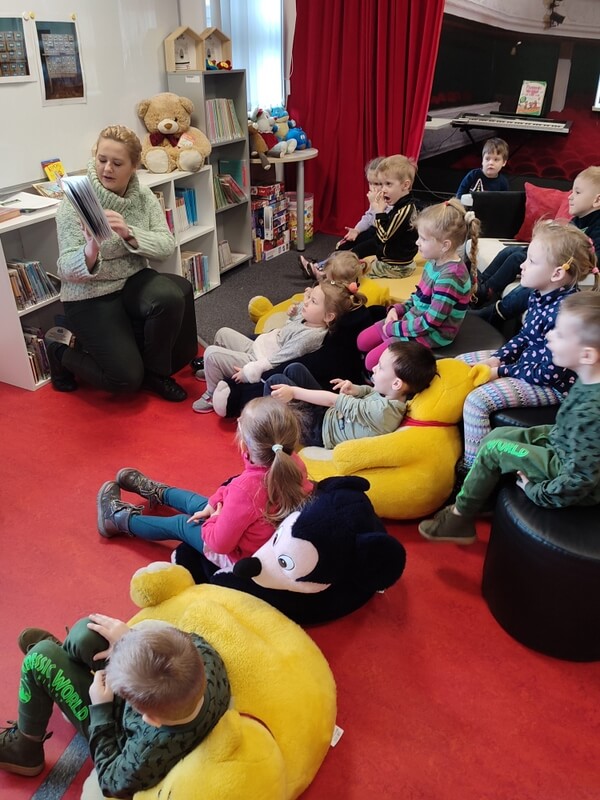  Po lewej stronie prowadząca zajęcia czyta dzieciom książkę. Po prawej stronie grupa dzieci skierowana twarzami do czytającej siedzi na siedziskach. Tło stanowi wystrój biblioteki.