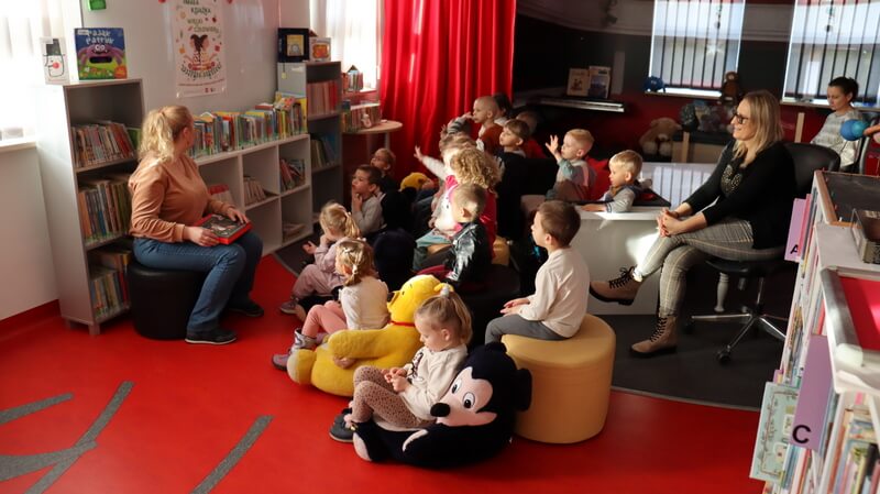 Po lewej stronie siedzi bibliotekarka. Z prawej siedzi grupa dzieci wraz z paniami, skierowana twarzami do prowadzącej. Tło stanowi wystrój biblioteki w postaci regałów z książkami.