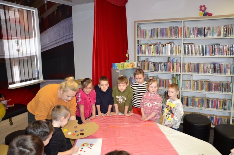 Grupa dzieci stoi wraz z prowadzącą wokół stołu na którym tworzą wspólny obraz. Tło stanowi wystrój biblioteki i regały z książkami.