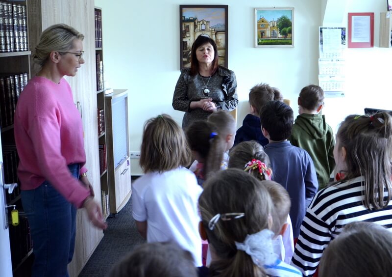 Grupa dzieci wraz z wychowawczynią odwiedzają Panią Dyrektor - Miejskiej Biblioteki Publicznej we Włodawie w jej gabinecie. Tło stanowią meble z książkami.
