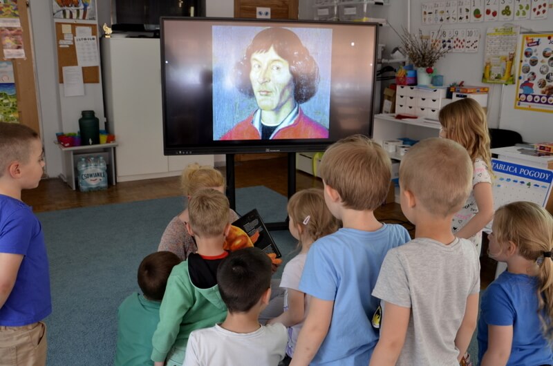 Grupa dzieci stoi przed prowadzącą, która pokazuje im kolorowe plansze z planetami. Za prowadzącą widać portret Mikołaja Kopernika. Tło stanowi wystrój sali przedszkolnej. 