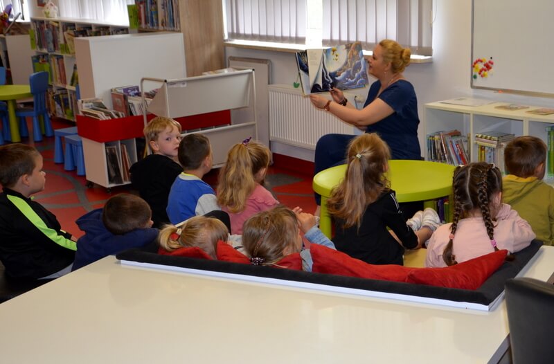 W oddali prowadząca czyta, pokazując dzieciom ilustracje. Przed nią siedzi grupa dzieci skierowana twarzami do prowadzącej. Tło stanowi wystrój biblioteki czyli regały z książkami.