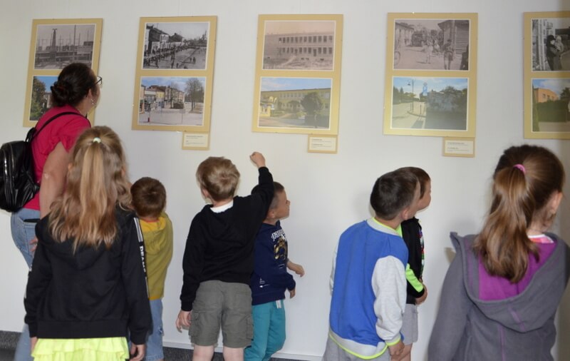 Grupa dzieci wraz z opiekunem ogląda wystawę. Skierowani są twarzami do wyeksponowanych fotografii na białej ścianie. 