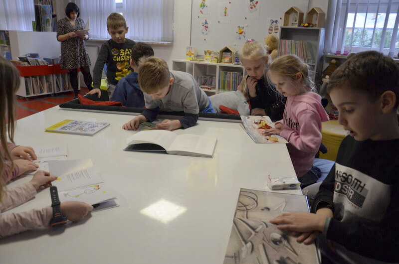 Oddział dla Dzieci. Przy białym stole w kształcie fortepianu siedzą i stoją dzieci, które oglądają kolorowe książki w języku ukraińskim. Próbują czytać, porównują literki z książek polskich. W tle na ścianie wisi biała tablica magnetyczna, na której przyczepione są kolorowe obrazki z motywem zimowym. Poniżej widoczny jest biały regalik z barwnymi książeczkami dla dzieci. Po lewej stronie, w tle stoi nauczycielka i przegląda komiks. Poniżej stoją czerwono białe regały z komiksami dla dzieci i młodzieży. 