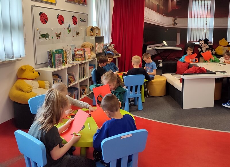 Dzieci w grupach siedzą przy stolikach. Podczas warsztatów wykonują swoje prace. Tło stanowi wystrój biblioteki w kolorach czarno- czerwonych. Z lewej strony stoją regały z książkami oraz dekoracje ścienne.