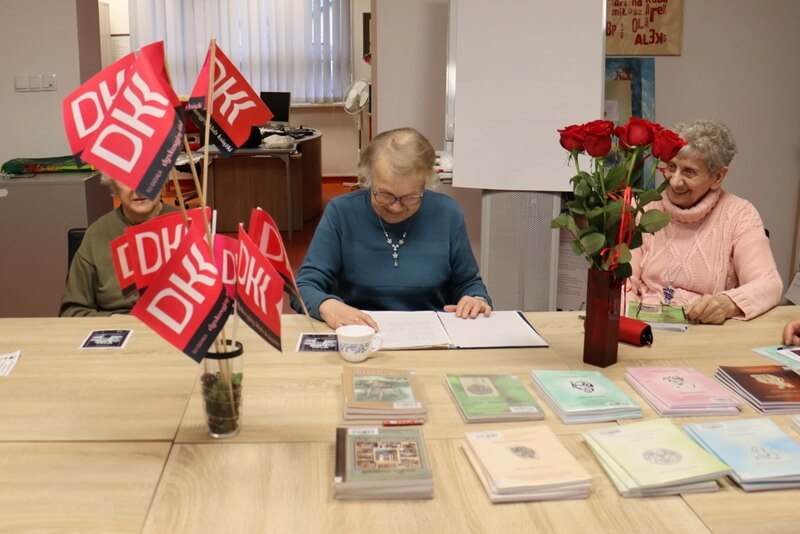 Trzy kobiety siedzą przy stole, przed nimi leżą książki oraz stoją dwa wazony - w jednym z nich znajdują się kwiaty, w drugim chorągiewki z napisem DKK