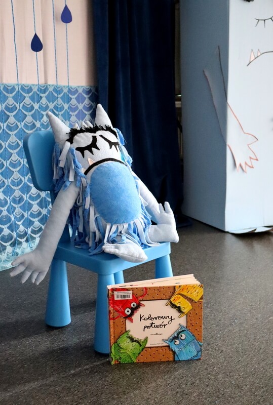 Na pierwszym planie jest wyeksponowana książka Kolorowy potwór. Na niebieskim krzesełku siedzi niebieski pluszowy potwór. Tło stanowią biało- niebieskie dekoracje.