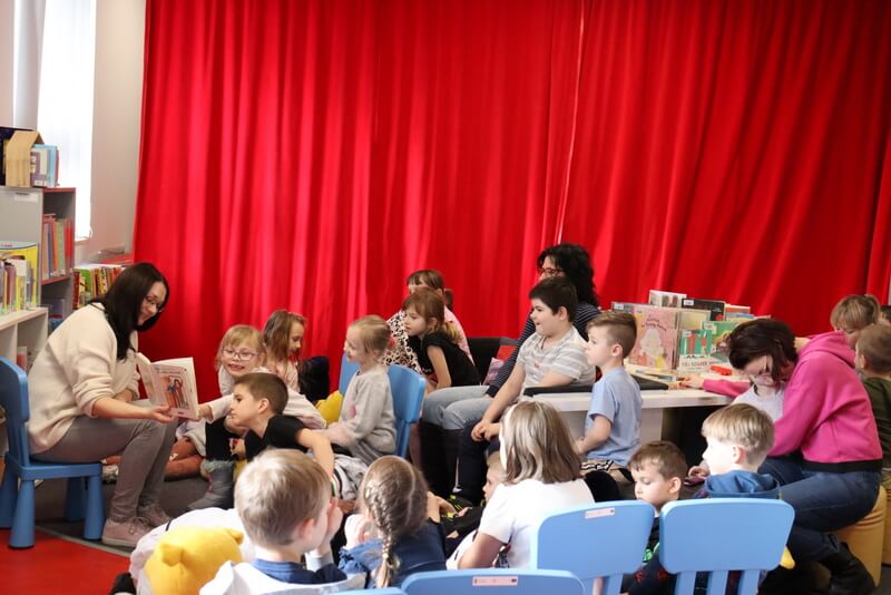 Oddział dla Dzieci. Grupa dzieci siedzi przed prowadzącą zajęcia (J. Orzeszko), która pokazuje im ilustracje czytanej książki. Część dzieci siedzi na kanapie w kształcie fortepianu, inne siedzą na niebieskich krzesełkach, jeszcze inne na pluszowych siedziskach. W tle wisi soczyście czerwona zasłonięta kurtyna. Na białym blacie stołu fortepianowego stoją książki dla dzieci, przygotowane do oglądania.