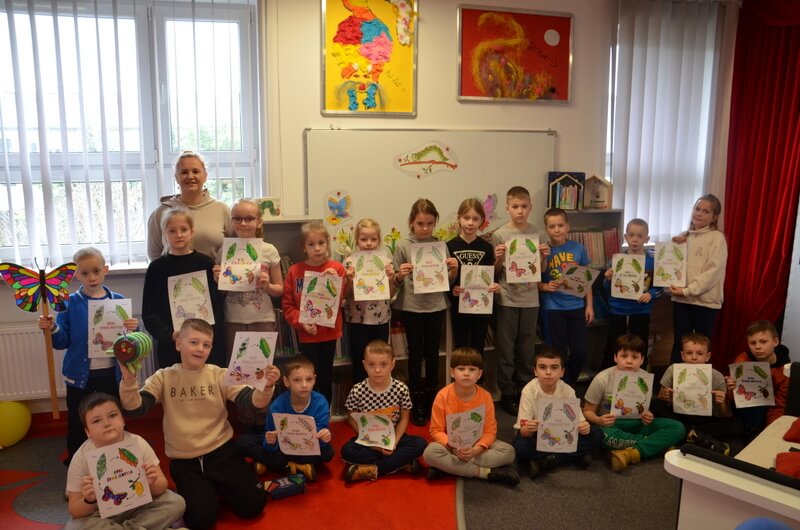 Grupa dzieci wraz z bibliotekarką pozują do wspólnej fotografii. Dzieci trzymają w rękach swoje prace.