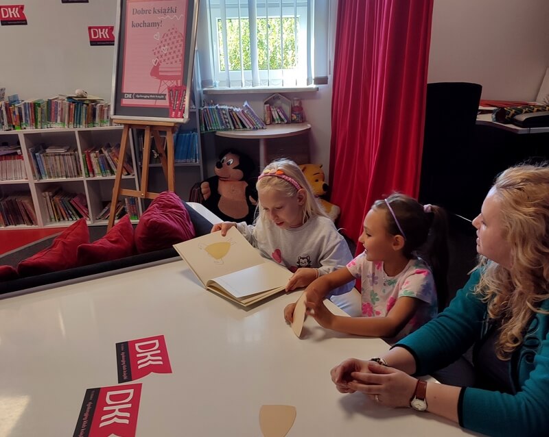 Jedna z dziewczynek czyta książkę wśród klubowiczów i bibliotekarki. W tle znajdują się książki oraz materiały promujące DKK.