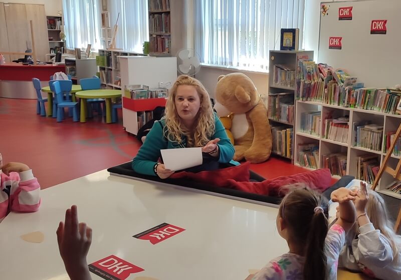 Grupa klubowiczów zgłasza się do odpowiedzi. W oddali siedzi bibliotekarka. Tło stanowi wystrój biblioteki w postaci regałów z książkami.