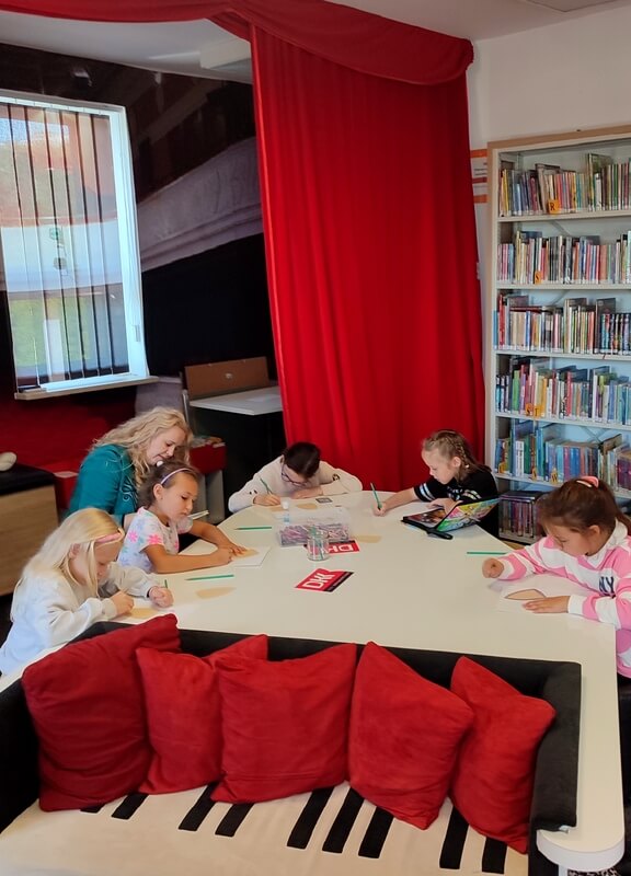 Grupa dzieci wraz z bibliotekarką tworzą prace. Tło stanowi wystrój biblioteki w postaci regałów z książkami.