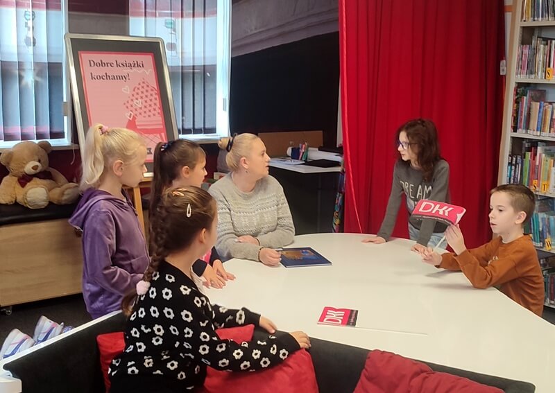 Grupa dzieci wraz z bibliotekarką siedzą wokół stołu, na której leży wyeksponowana książka i chorągiewki DKK. W oddali widnieje wyeksponowany plakat DKK. Tło stanowi wystrój w postaci regału z książkami.