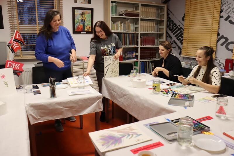 Czytelnia Działu Regionalnego. Prowadząca warsztaty (niebieski sweter) i moderator Dyskusyjnego Klubu Książki (szara koszulka) otwierają zajęcia. Moderator J. Orzeszko pokazuje blok bambusowy młodym kobietom siedzącym przy stole. Stół nakryty białym, papierowym obrusem. Na stole przed każdą uczestniczką stoi słoik z wodą, pędzle, kolorowe farby i papier do malowania. W tle widoczne różowe chorągiewki z logo Dyskusyjnego Klubu Książki DKK, regały z książkami.