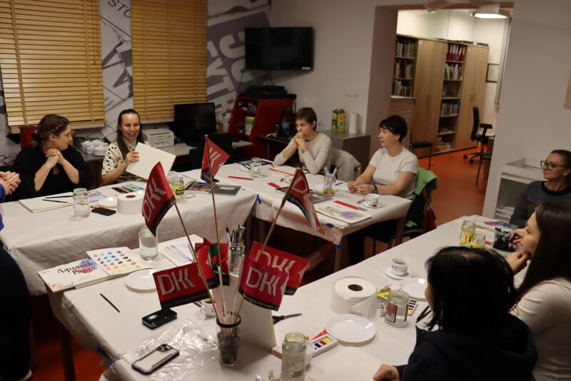 Czytelnia Działu Regionalnego. Stoły przykryte białymi obrusami papierowymi. Na stołach kolorowe farby, kartki papieru i rysunki. Przy stołach siedzą uśmiechnięte młode kobiety, oglądają farby, pędzle. Na pierwszym planie różowe chorągiewki z logo Dyskusyjnego Klubu Książki. U góry w rogu Czytelni widać wyłączony czarny telewizor. W oknach drewniane rolety.