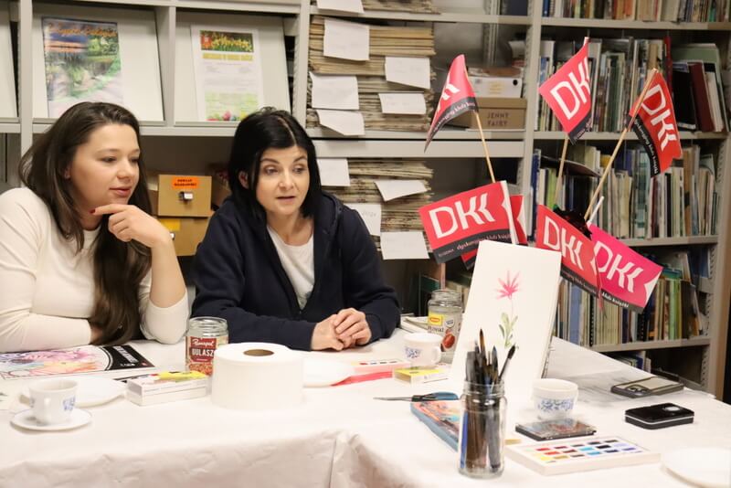 Dwie uczestniczki zajęć rozmawiają o obrazku, na który patrzą. Obrazek oparty jest o flakon z różowymi chorągiewkami DKK. Przed kobietami na stole leżą kolorowe farby, kartki, rysunki. Słoiki z wodą i pędzle.