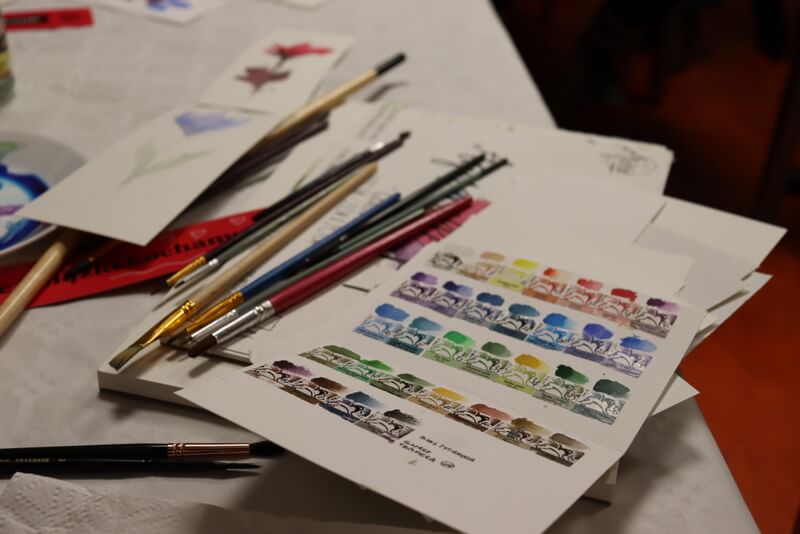 Zbliżenie na kolorowy kartonik z próbkami barw akwarelowych. Pędzle, papier i farbki. Widać też kolorowe, delikatne zakładki z kwiatkami.