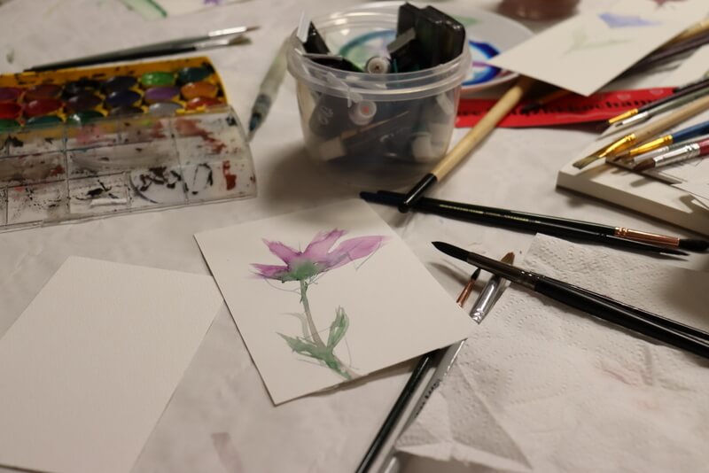 Zbliżenie na zakładkę z kwiatkiem. Rysunek delikatny, barwy fioletowo niebieskie. Obok pędzle, różne farbki w kostkach, w tubkach.