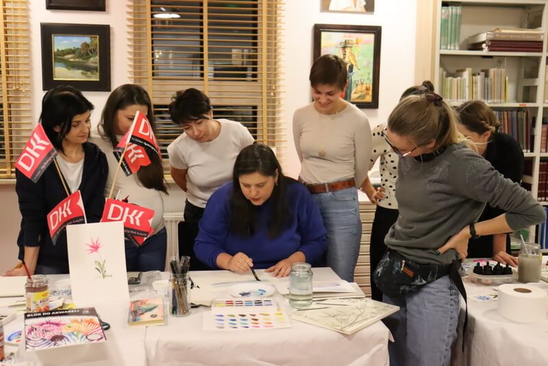 Czytelnia Działu Regionalnego. Stoły przykryte białymi obrusami papierowymi. Na stołach kolorowe farby, kartki papieru i rysunki. Kobiety zgromadzone dookoła prowadzącej warsztaty patrzą i słuchają, jak K. Melcher rysuje instruktażowy obraz. Młode kobiety zgromadzone wokół prowadzącej zainteresowane słuchają.