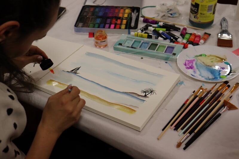 Ujęcie przez ramię malującej. Zbliżenie n piękny pejzaż nadmorski. Barwy miłe, ciepłe. Obok obrazu leżą farby i pędzle. Paleta do mieszania kolorów artystycznie przybrudzona.