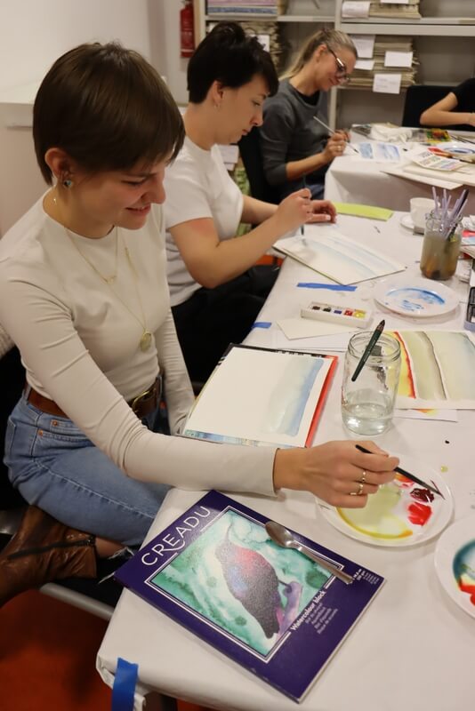 Uśmiechnięta uczestniczka zajęć maluje swój pejzaż, w tle widoczne pozostałe młode kobiety malujące w skupieniu. Na stołach pełno kolorowych farb, pędzli, słoiki z wodą. Blok z papierem do malowania akwarelą.